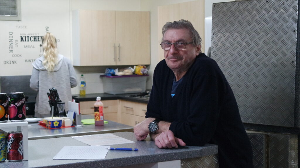 Man sitting at table in a community kitchen, woman at sink in background
